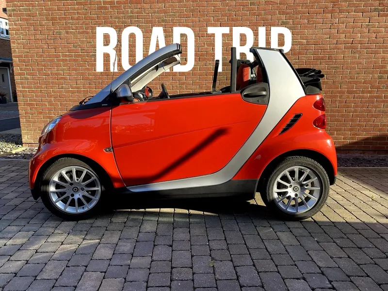 Red Smart car with ROAD TRIP in white Bebas Neue letters behind it, in front of a brick wall